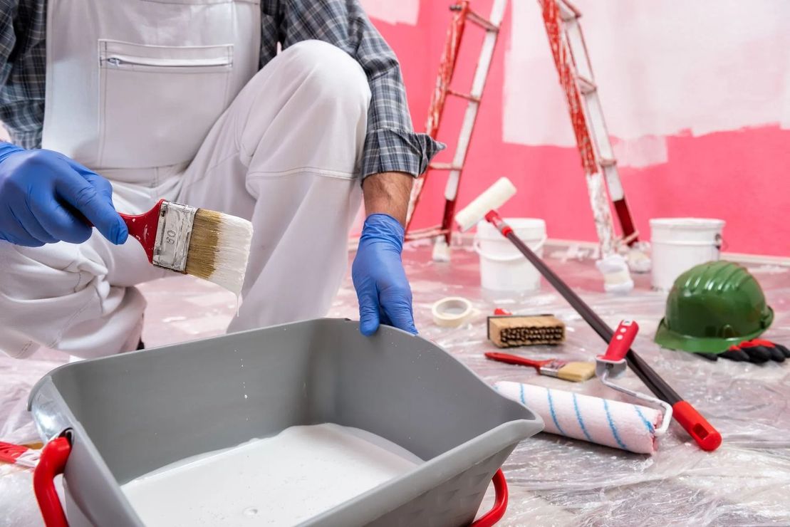 Painter in white overalls and blue gloves painting a room with a brush and tray near ladders and tools