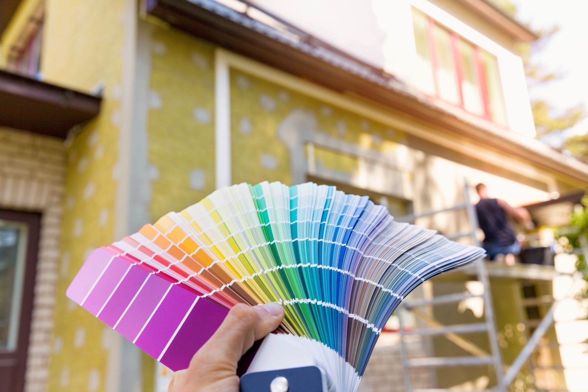 Hand holding a fan of colorful paint swatches outside a yellow house under construction