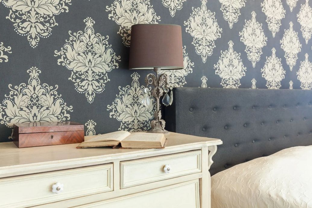 Gray tufted headboard beside a white dresser with a lamp against patterned wallpaper