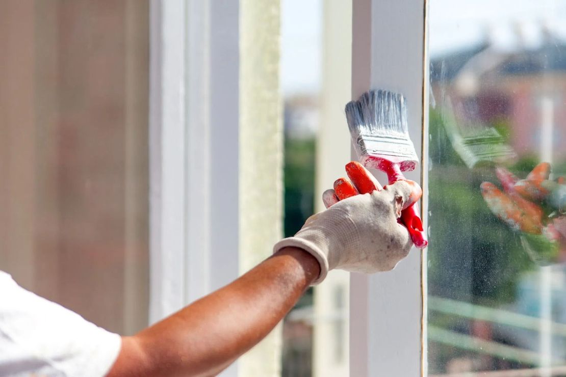 Gloved hand painting a white window frame with a brush near a glass pane