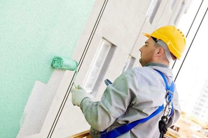 Painter applying green paint to a white wall while wearing a yellow hard hat and safety harness