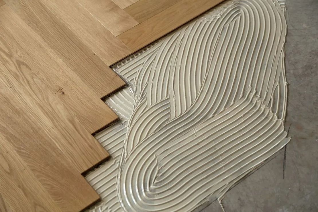 Wooden floor planks meeting gray adhesive spread with a notched trowel pattern