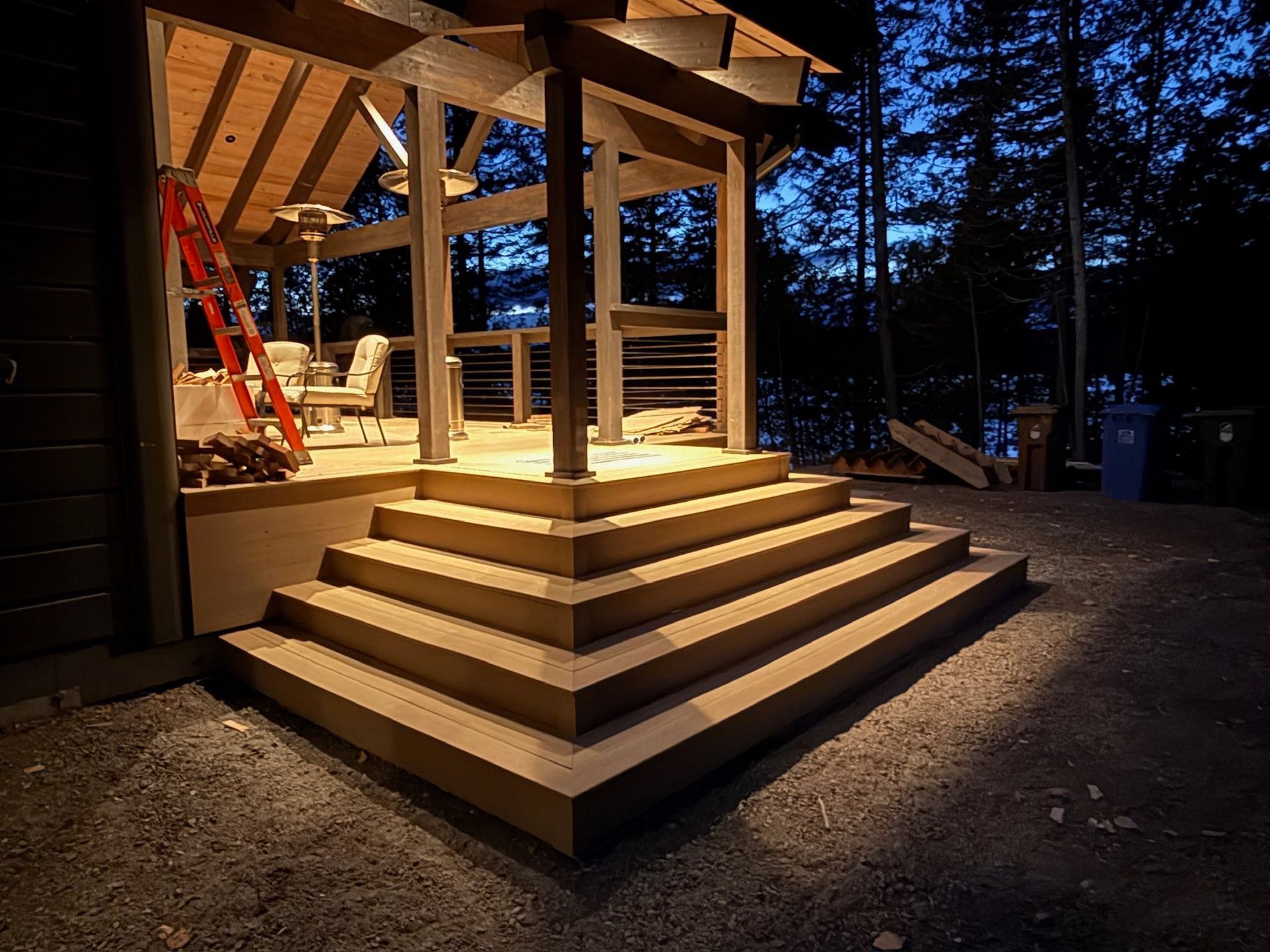 Au crépuscule, une entrée en terrasse de bois à plusieurs niveaux, éclairée, avec une échelle rouge et du mobilier de jardin visibles sur le porche.