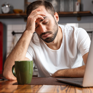 A man with a beard with sitting by his laptop, resting his head in his hand and trying not to fall asleep. His coffee sits next to his laptop ignored,