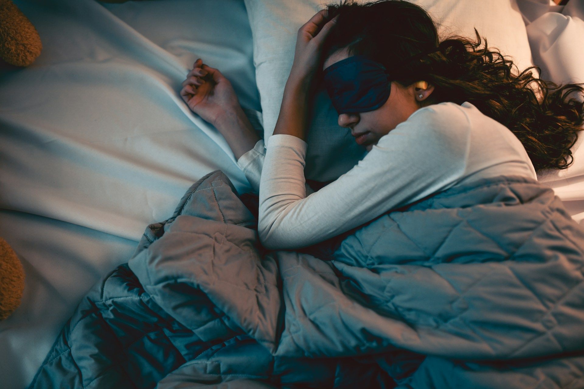 Person sleeping under blankets in a dimly lit bed, curled up with a pillow