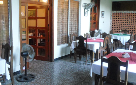 Comedor vacío de un restaurante con mesas vestidas con manteles rosas y blancos y un ventilador de pie junto a la puerta.