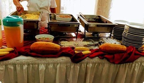 Mesa de buffet con bandejas cubiertas, platos, fruta y bebidas en un montaje para banquetes.