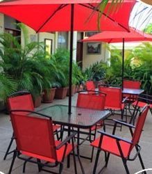 Patio exterior con sombrillas rojas, sillas rojas y mesas rodeadas de exuberantes plantas verdes.