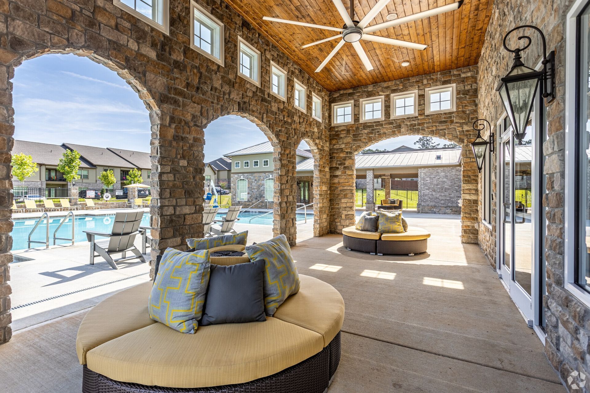 Stone arched patio overlooking a pool. Circular seating with pillows. Wooden ceiling, decorative lighting.