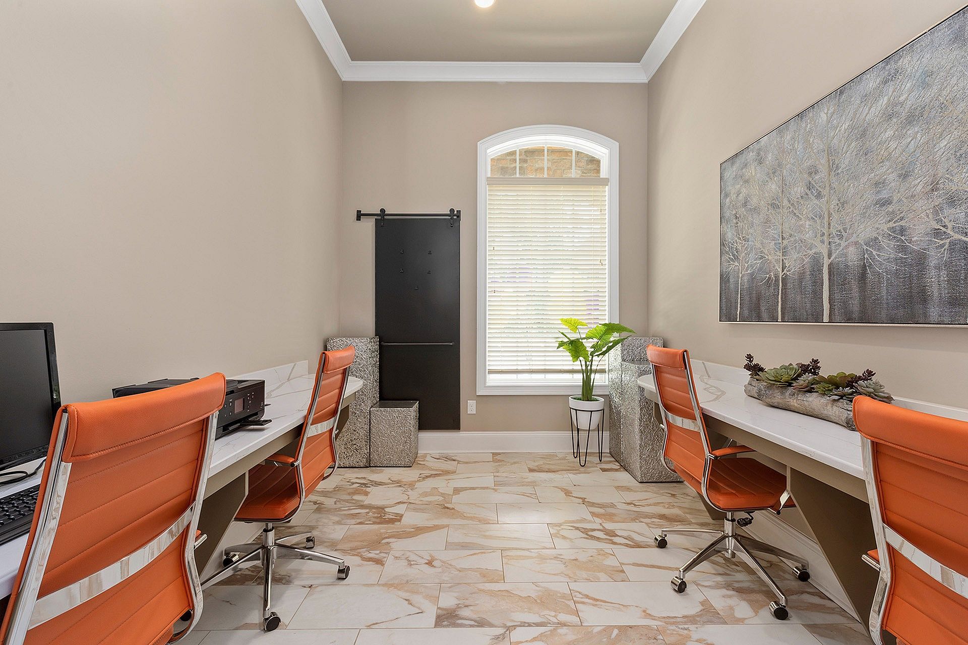 Community office space: two desks, orange chairs, window with blinds, plant, and artwork.