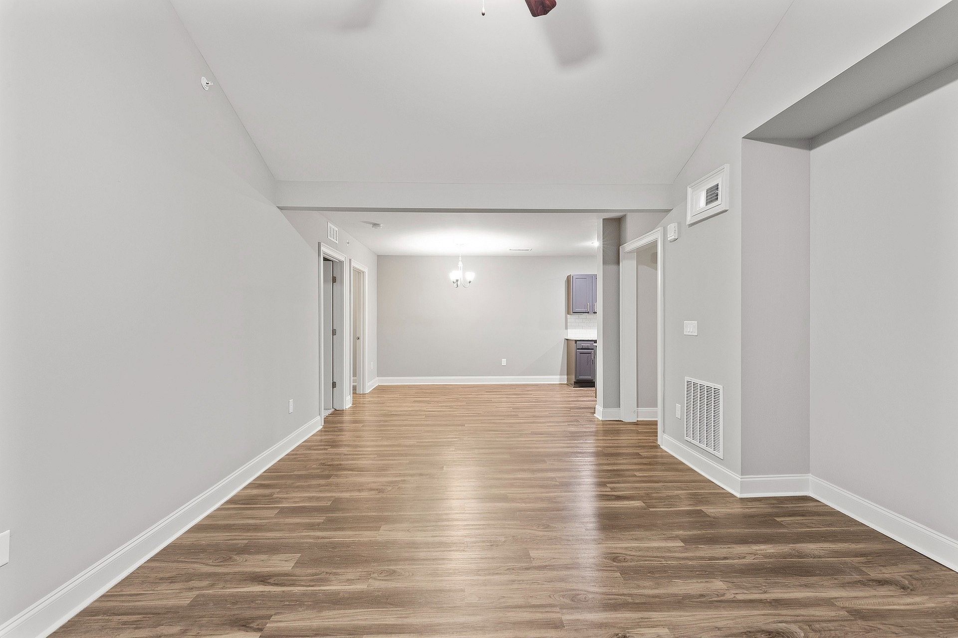 Empty living area of an apartment with wood flooring, gray walls, and open kitchen.