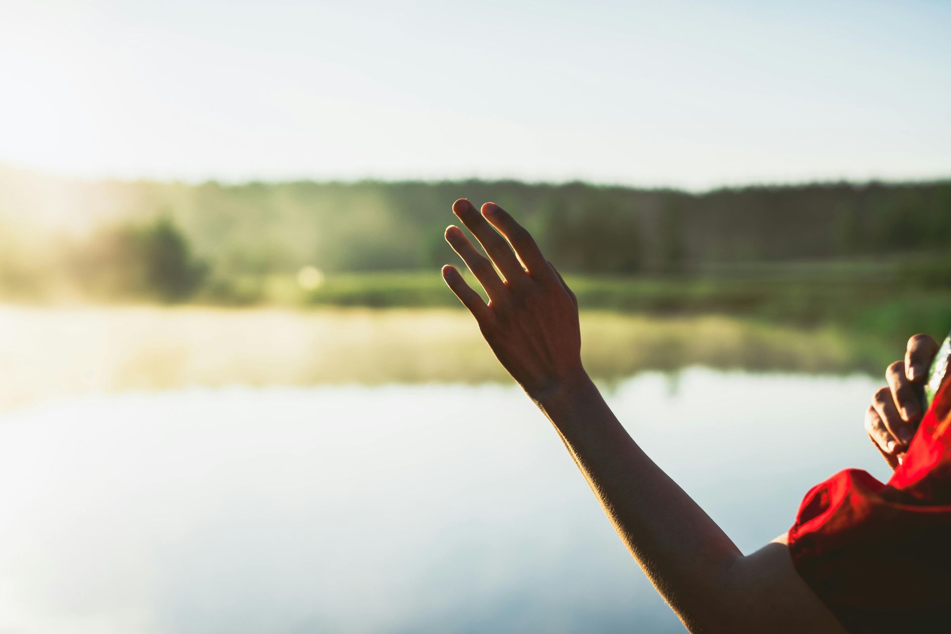 Raised hands in a red shirt by a sunlit lake with forest in the background