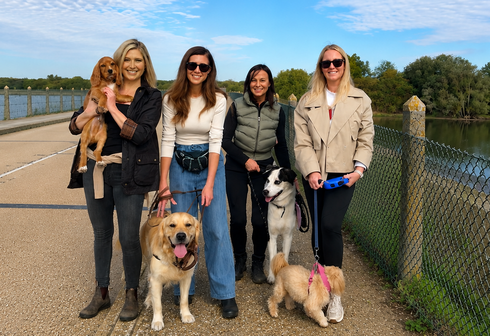 Four women with four dogs posing outdoors by a fence and lake on a sunny day.