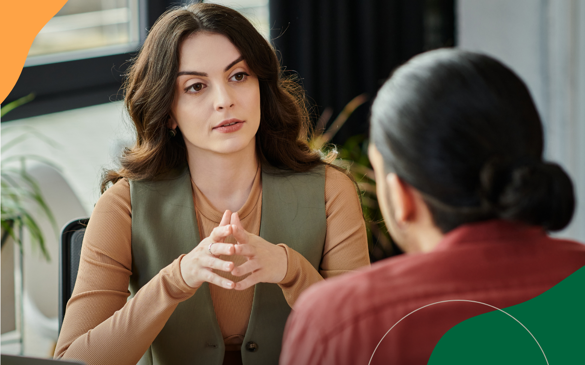 Two women talking across a table in a counseling session, one listening intently with hands clasped.