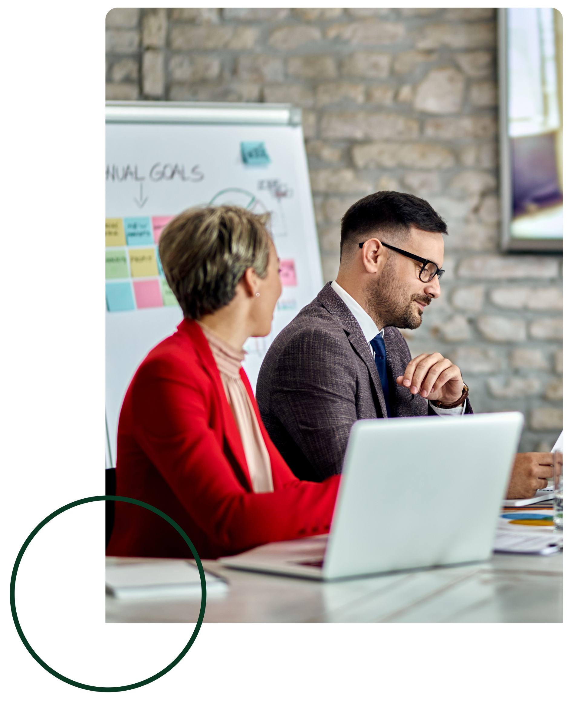 Two people in a meeting room with a laptop, whiteboard, and brick wall backdrop