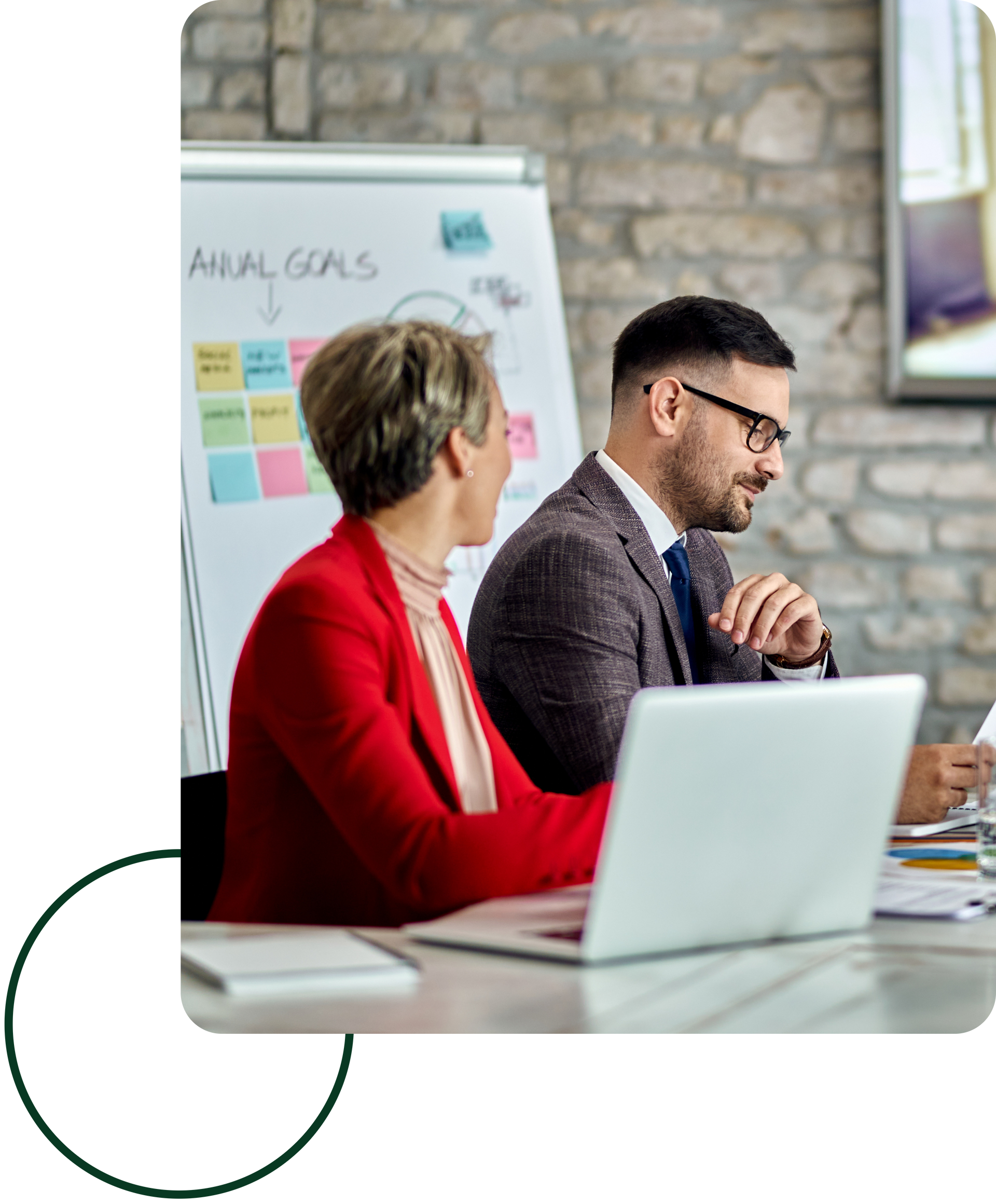 Two coworkers in a meeting beside a laptop and whiteboard with sticky notes in a brick-walled office