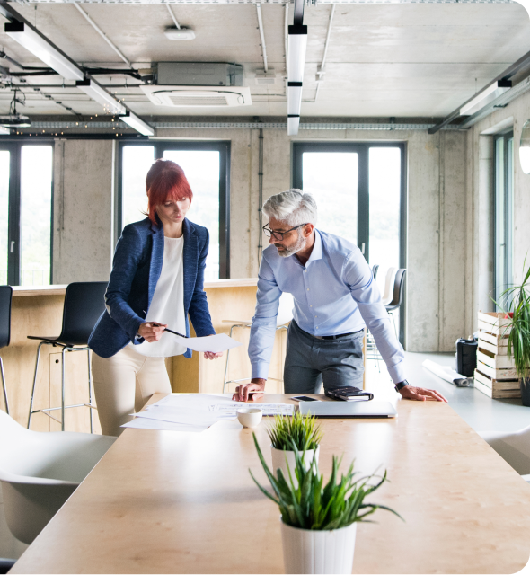 Two coworkers lean over a table in a bright modern office, discussing paperwork with a laptop nearby.