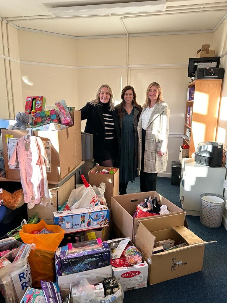Three women standing in a room filled with boxes, bags, and assorted items, with a large window on the right.