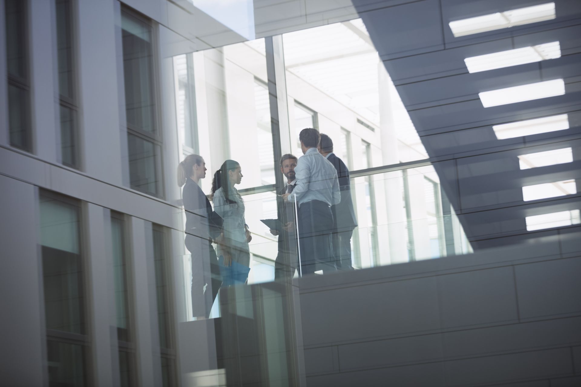 People standing in a bright glass hallway, seen through large windows.