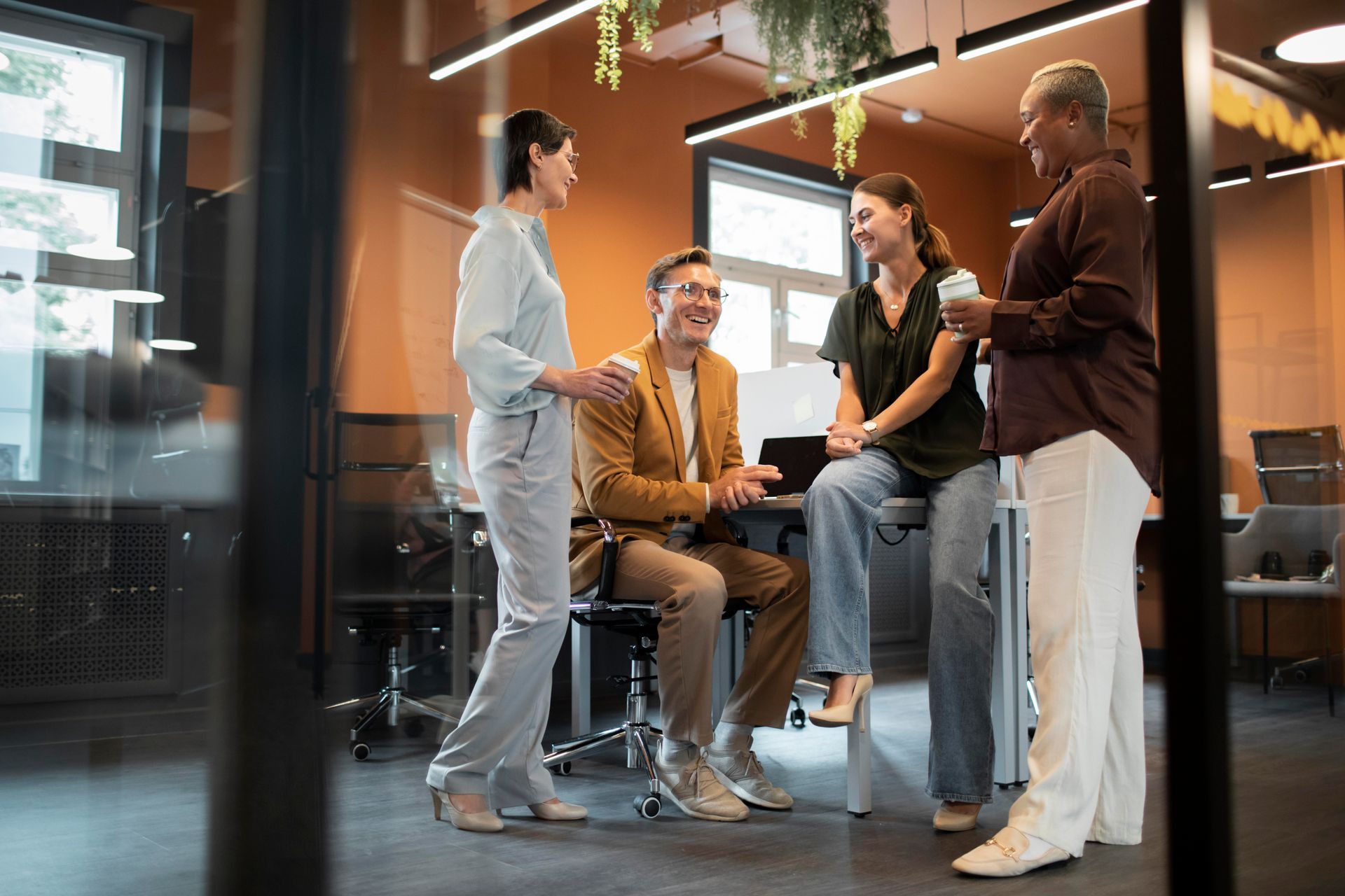 Four coworkers chatting in a modern office lobby, with one seated and others standing nearby.