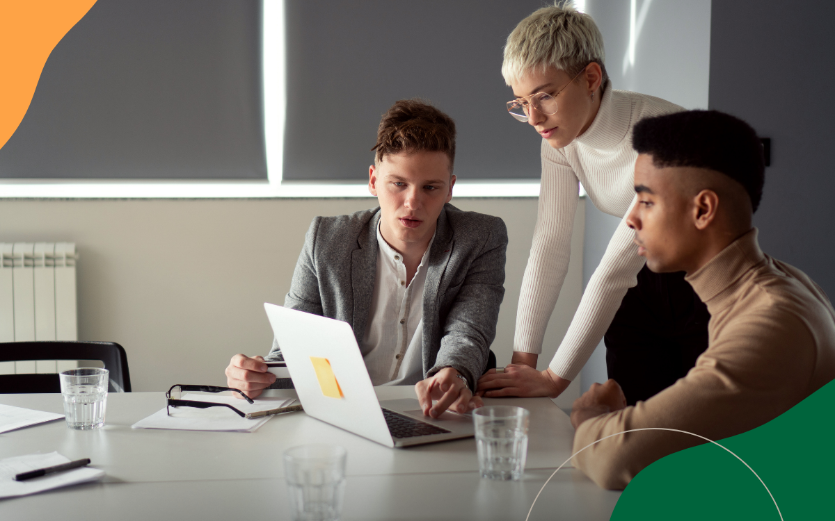 Three coworkers discuss a laptop at a conference table in a bright office meeting room.