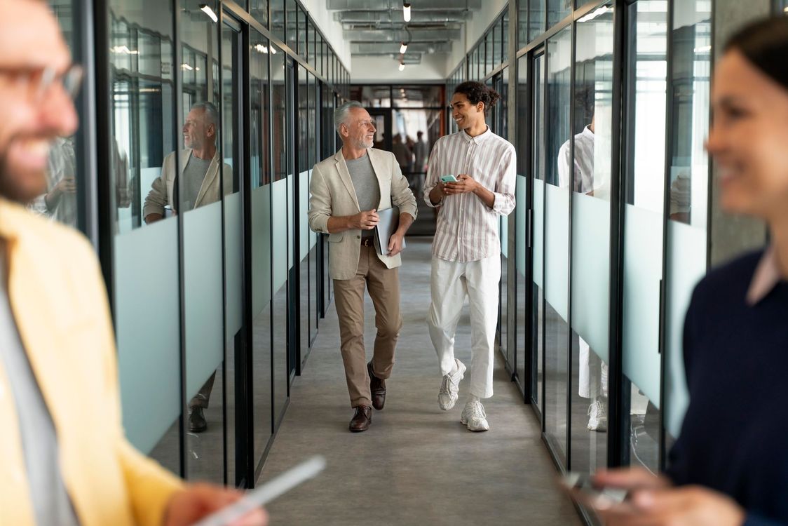 Two coworkers walking and talking in a glass-walled office hallway, holding papers.