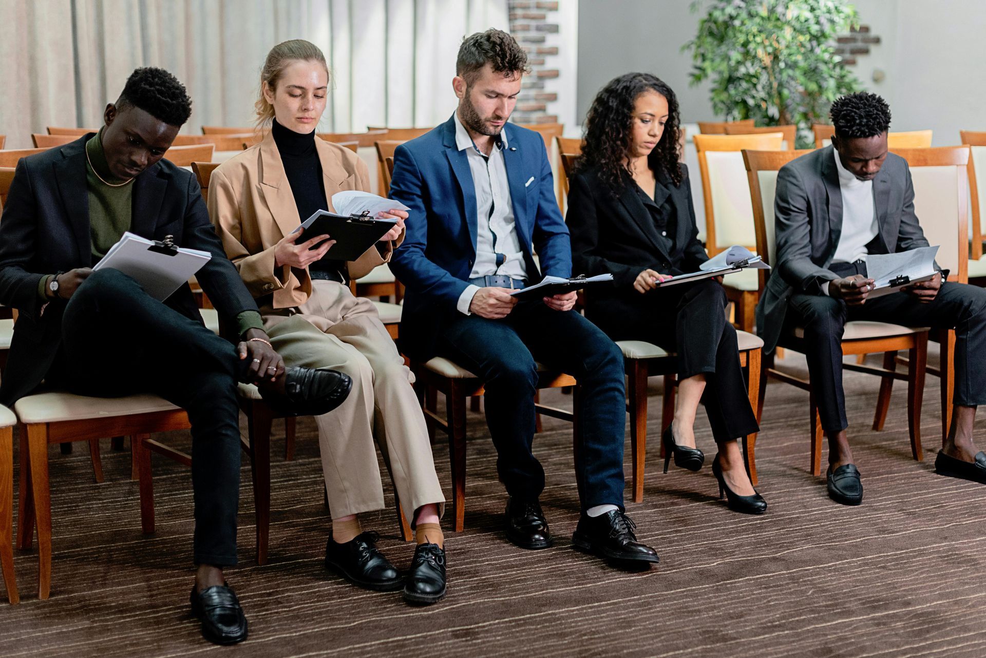 Five people in business attire sit in a row on chairs, each holding documents or clipboards while waiting.