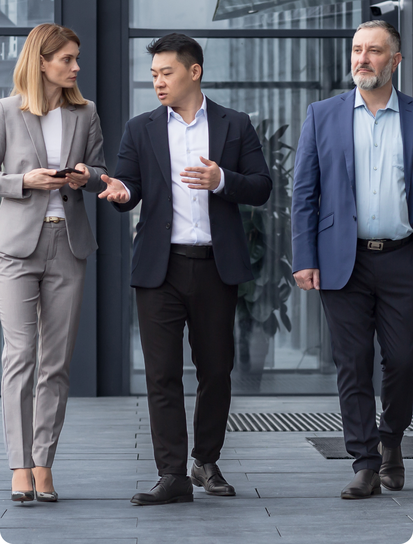 Three businesspeople walking outside a glass office building, talking together.