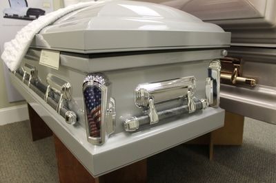 Gray casket with silver hardware, displayed indoors, with white fabric draped over the top.