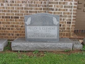 Gravestone of Peggy S. Clement with praying hands carved above her name and dates; set against a brick wall.