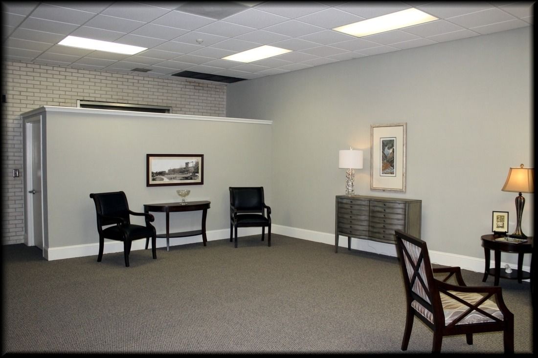 Empty waiting room with gray walls, chairs, small tables, and artwork. Fluorescent lighting illuminates the space.