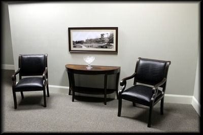 Two black chairs flank a dark wood table with a framed landscape. Gray walls and carpet.
