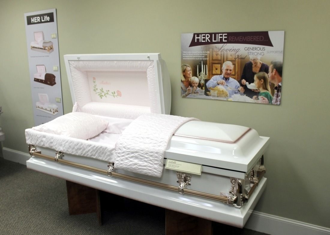 White casket, open, with pink bedding. Displayed on a platform with decorative trim, in a funeral home.