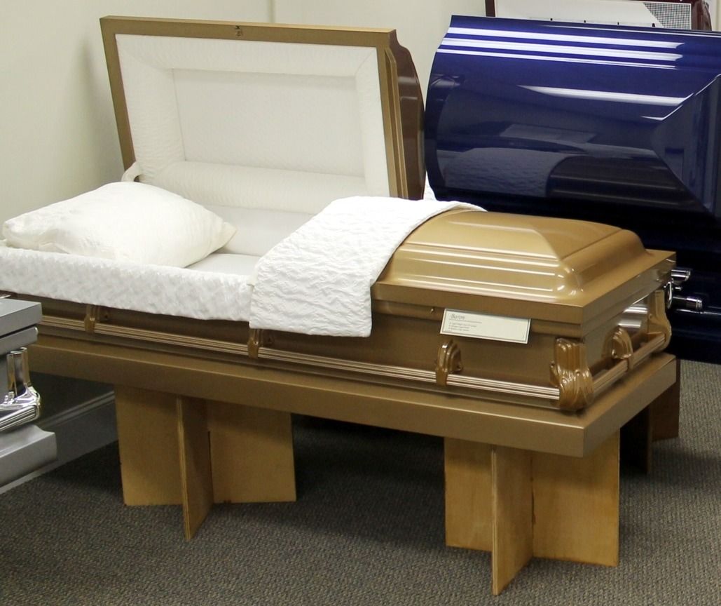 Gold coffin, open with white bedding, displayed on a wooden stand. A blue coffin is partially visible in the background.