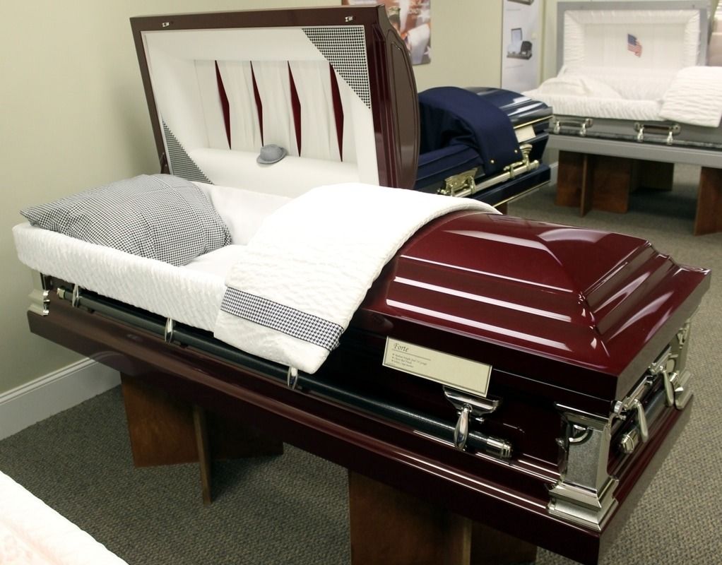 Maroon casket, partially open, with pillow and blanket inside. Other caskets in the background, a funeral home setting.