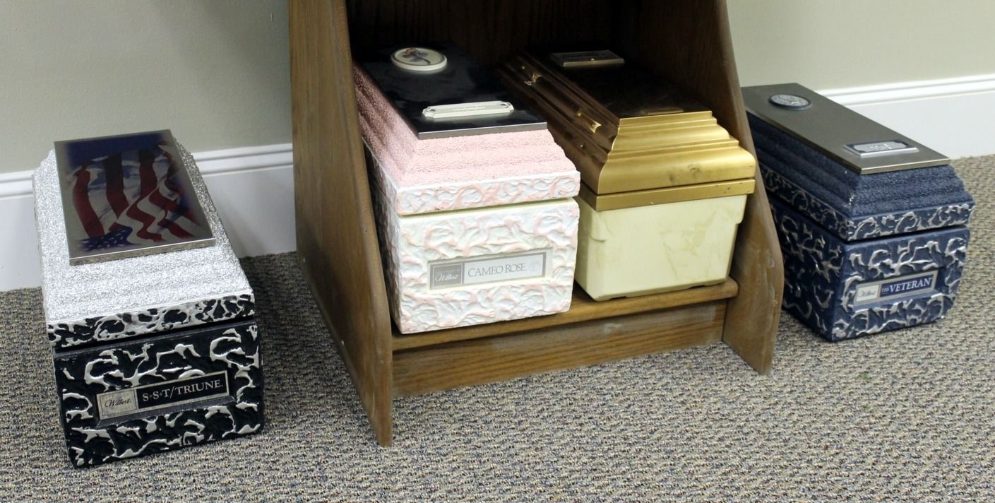 Cremation urns on display, various colors and styles, set on a wooden shelf and floor, near a wall.