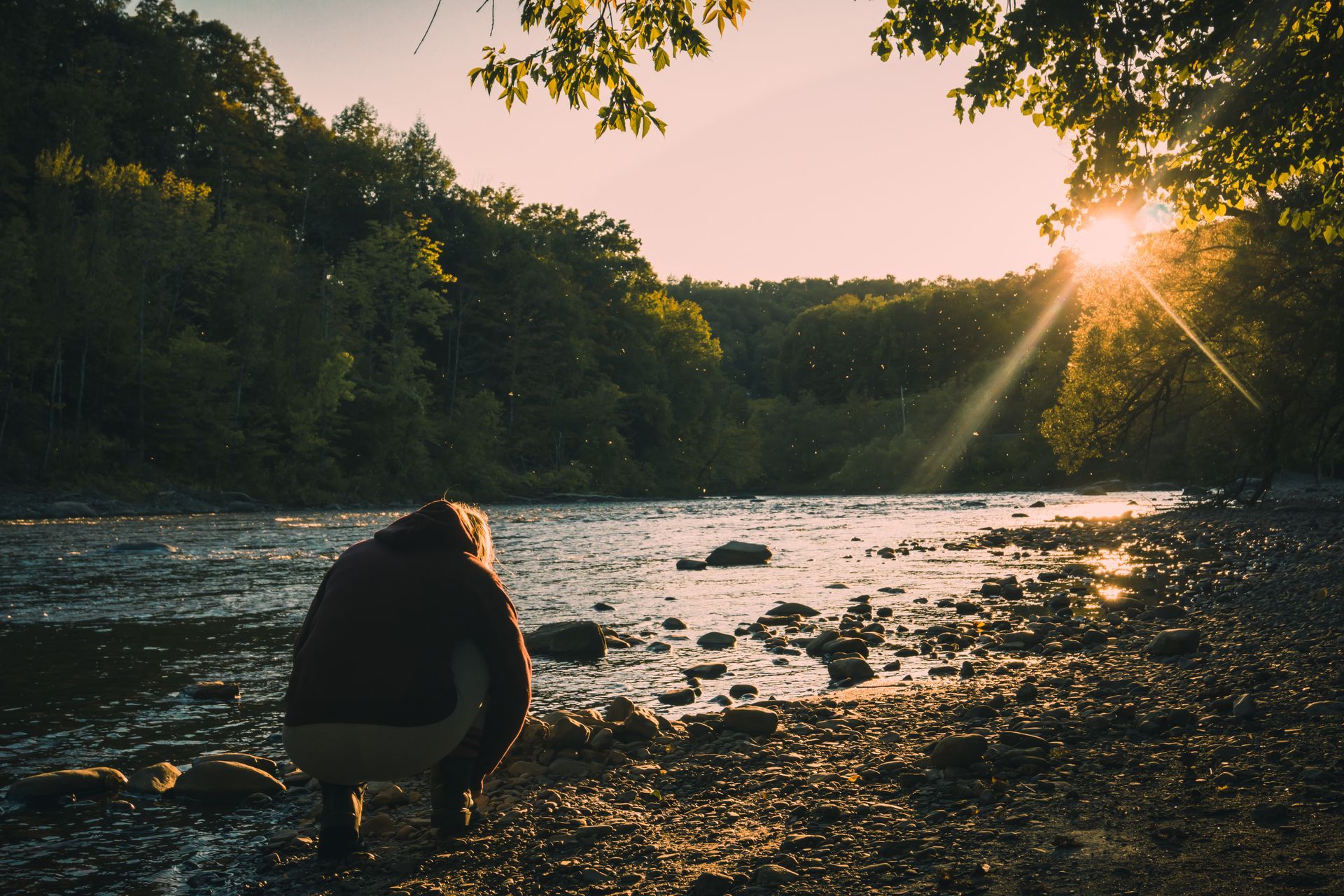 Person crouches at river's edge, gazing at the sun setting over forested hills.