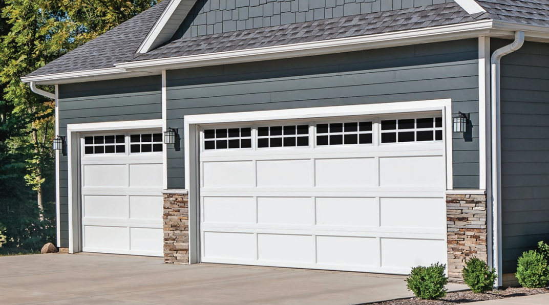 Two white garage doors with window panes on a blue-gray building with stone accents, concrete driveway, and green bushes.