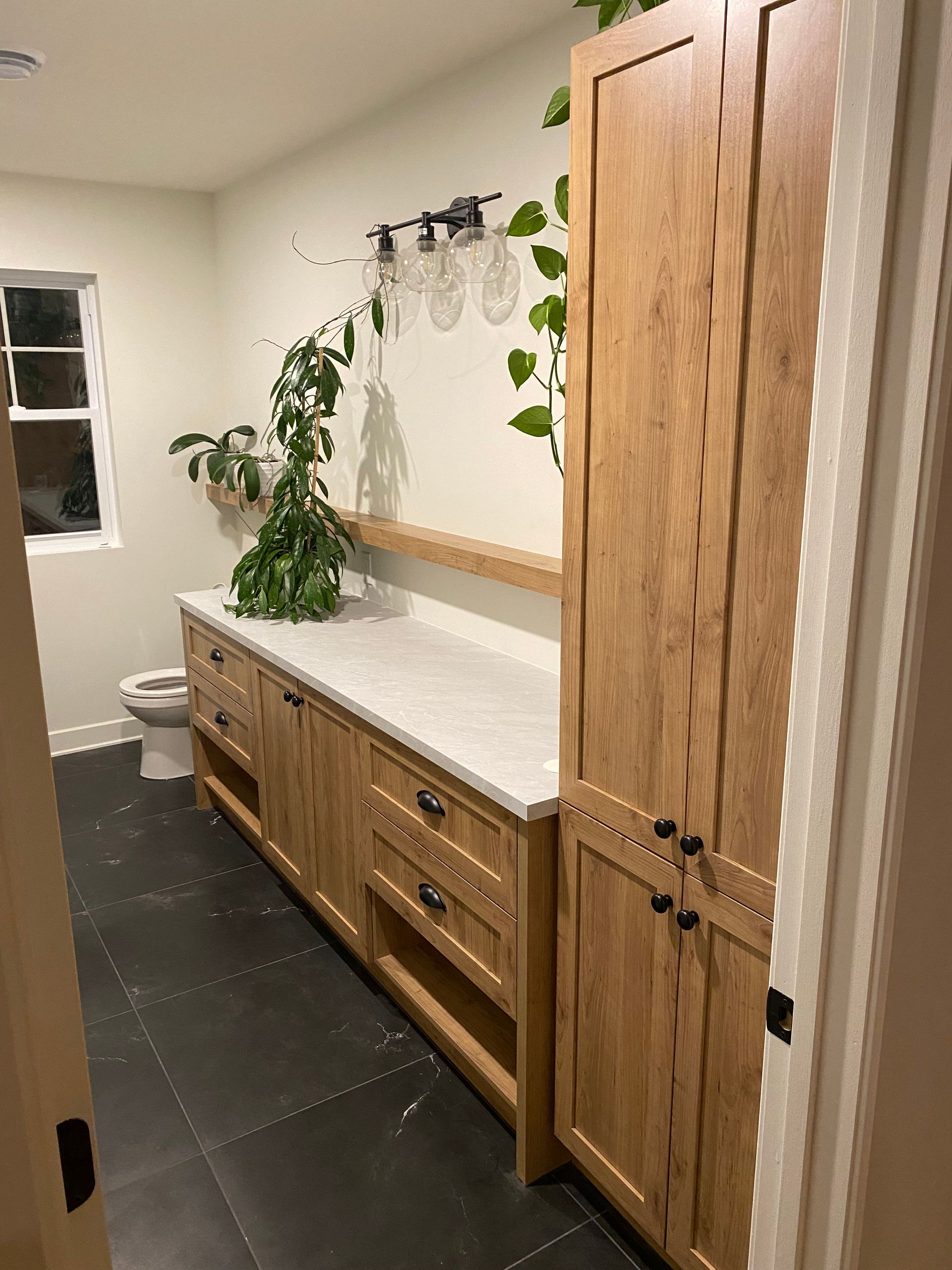 Salle de bains avec meuble-lavabo en bois, armoire et sol foncé ; des plantes décorent l'espace.