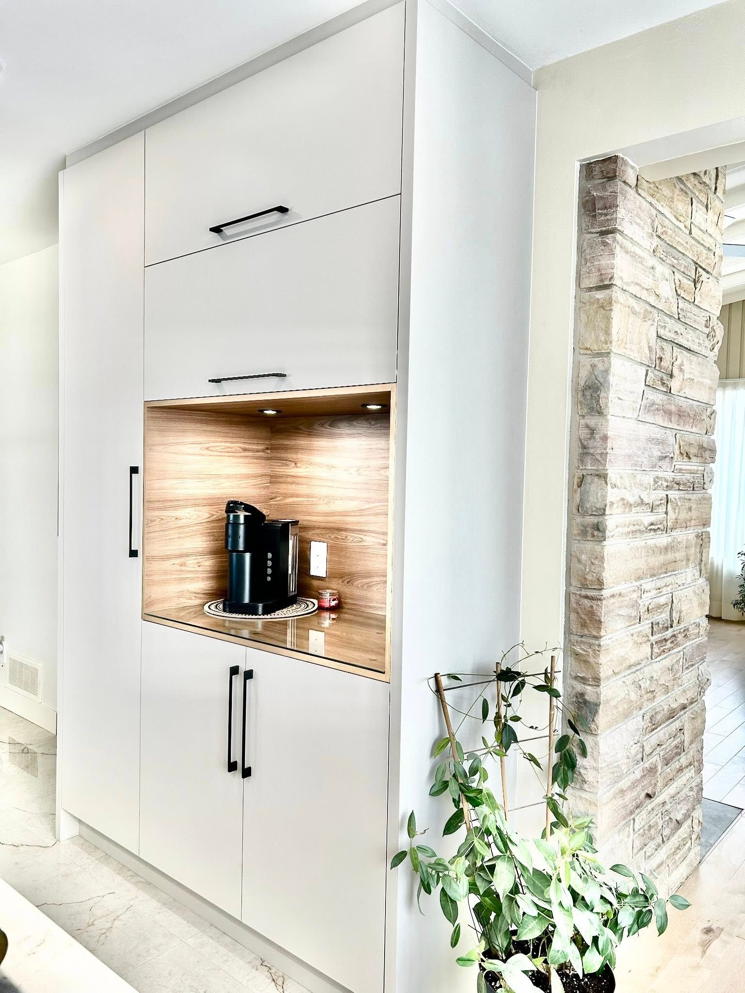 Meubles de cuisine blancs intégrés avec cafetière dans une niche en bois. Une plante en pot est visible au premier plan.