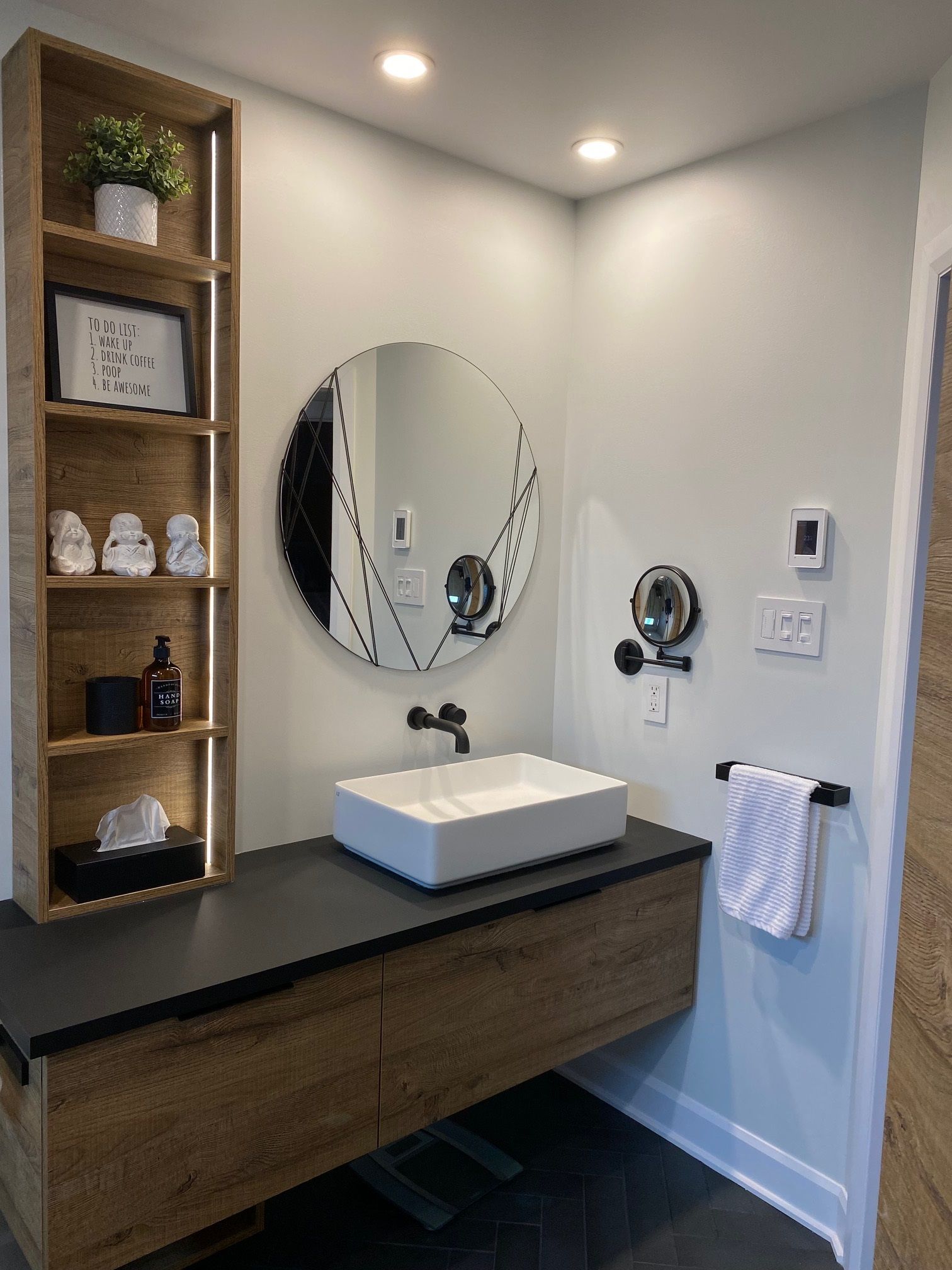 Salle de bains moderne avec meuble-lavabo suspendu, miroir rond et étagère en bois accueillant des objets de décoration.