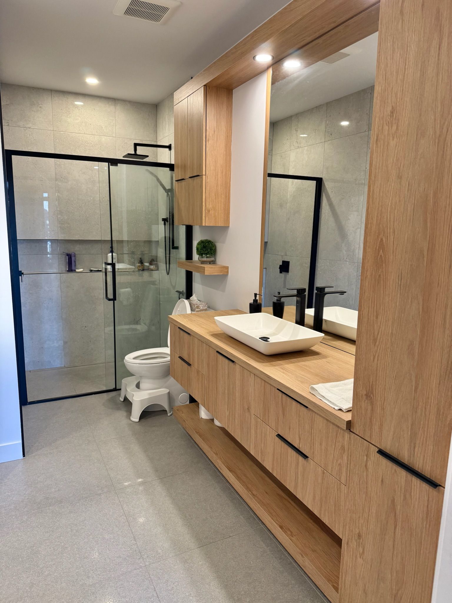 Salle de bains moderne avec meuble-lavabo en bois, grand miroir, douche en verre et sol gris.