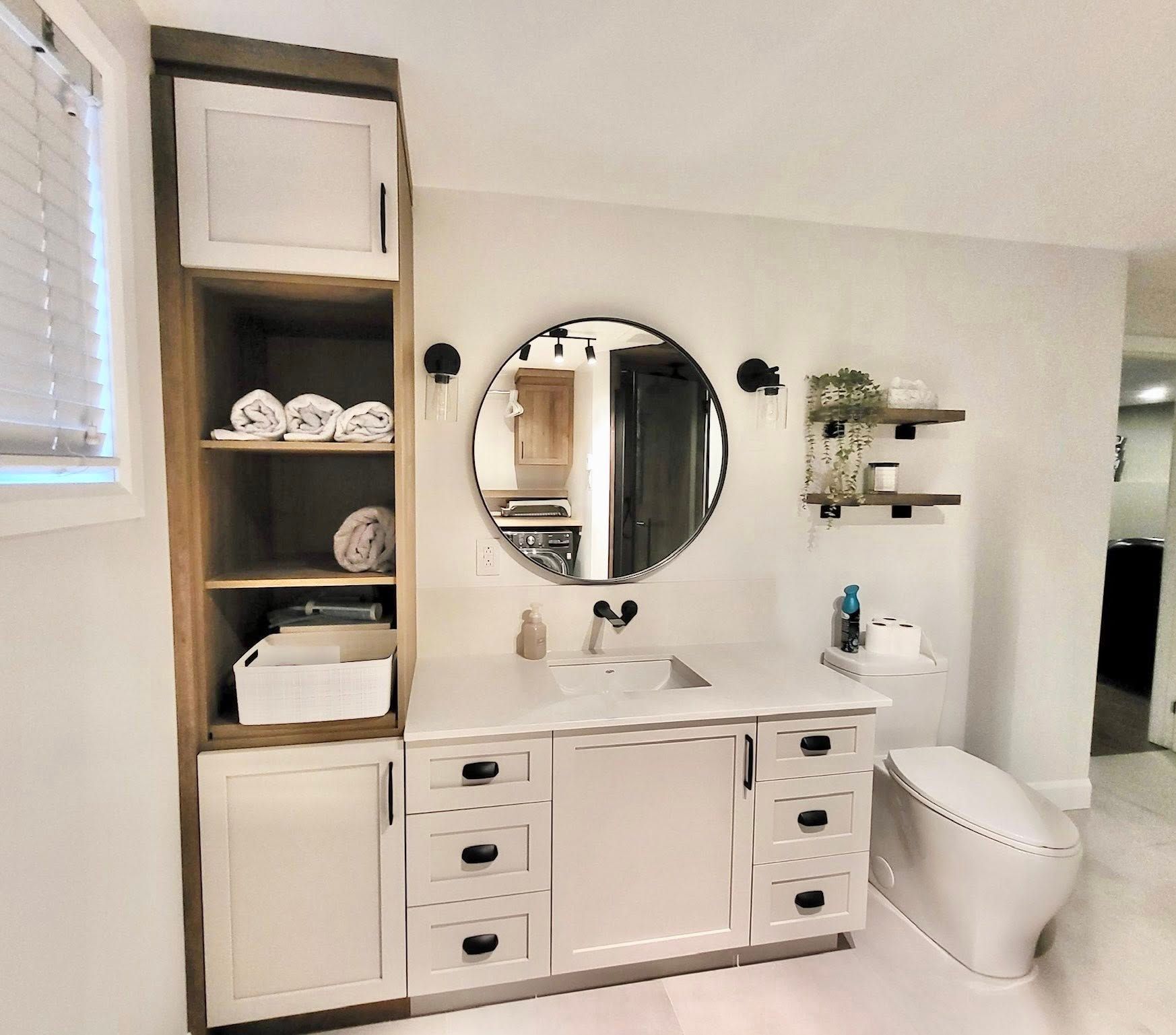 Salle de bains avec meuble-lavabo blanc, miroir rond, étagères et toilettes modernes.