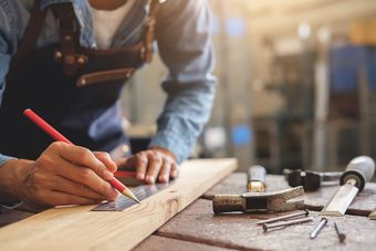 Personne dans un atelier utilisant un crayon et une règle pour marquer une planche de bois sur un établi, avec des outils.
