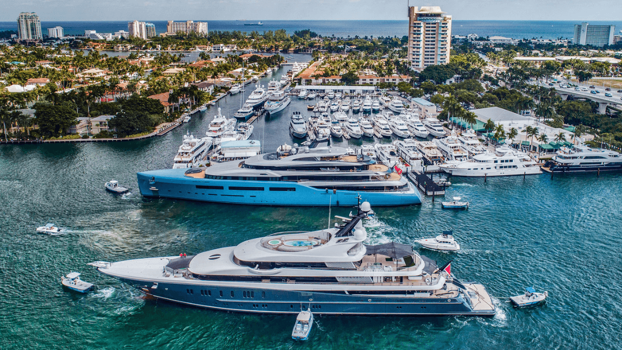 Aerial view: yachts docked in a harbor, turquoise water, boats nearby, buildings and trees in the background.