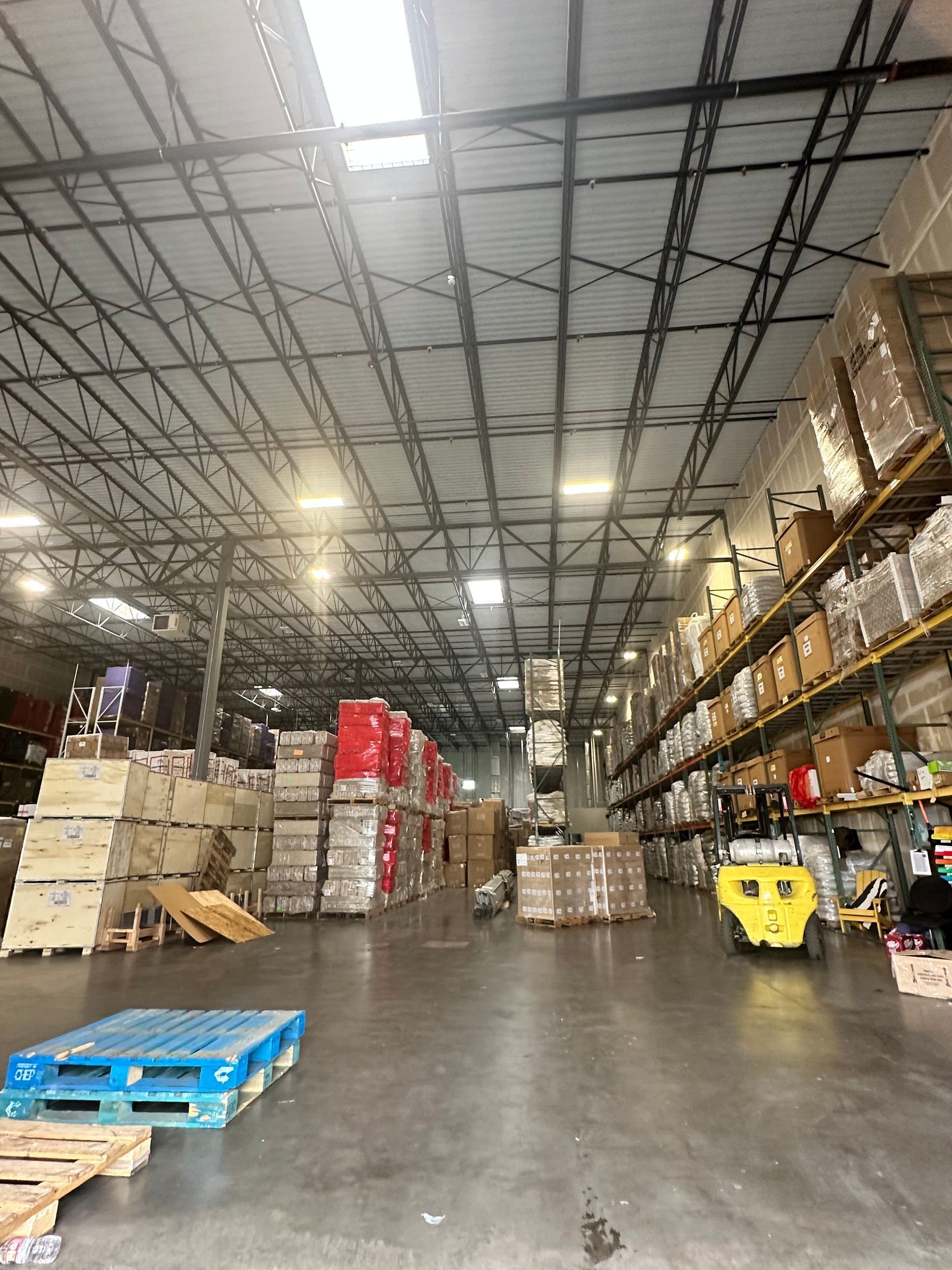 Warehouse interior with pallets, stacked boxes, and shelving. Skylights in the ceiling illuminate the space.