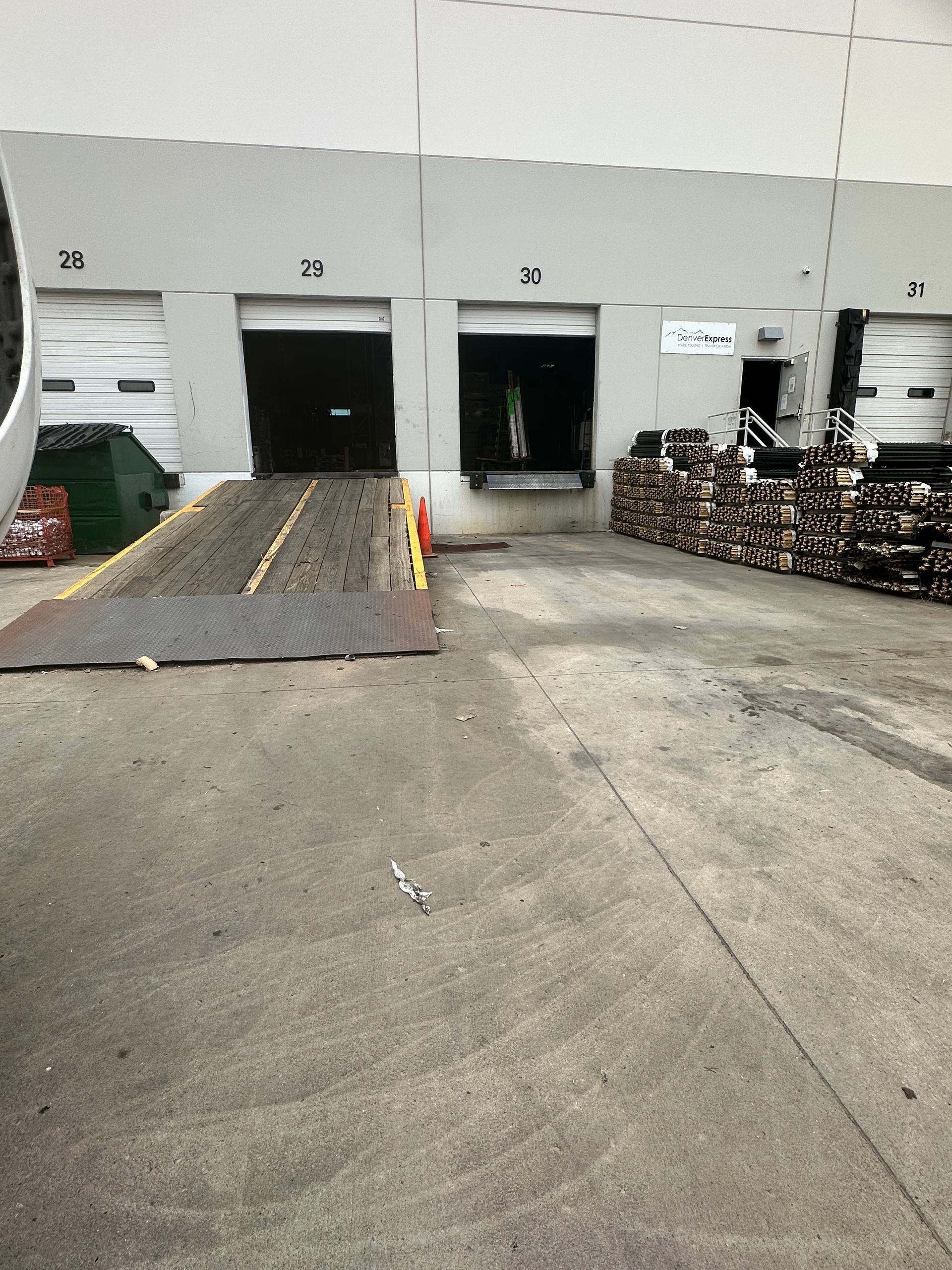 Loading dock with open bay doors. A ramp extends to the ground, with stacks of wood and a dumpster nearby.