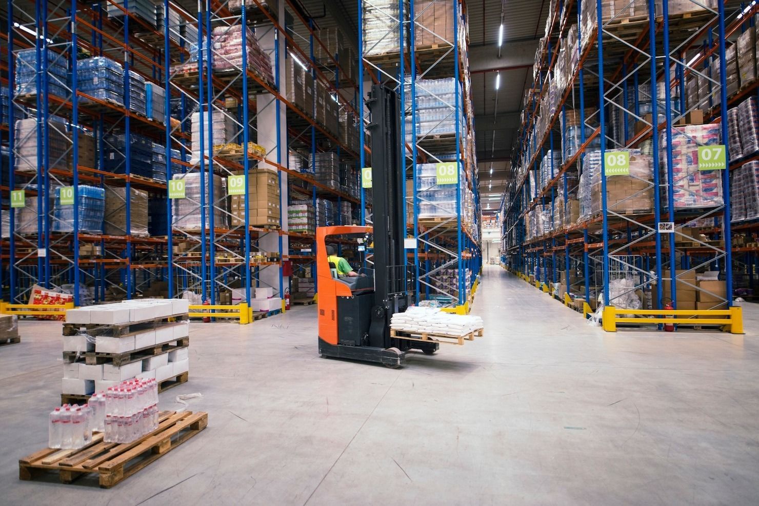 Forklift in warehouse picking up a pallet; shelves filled with stored boxes.