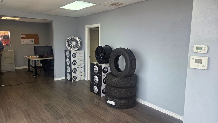 Empty auto shop waiting area with stacked tires, wheel displays, desks, and wall thermostat on gray walls.