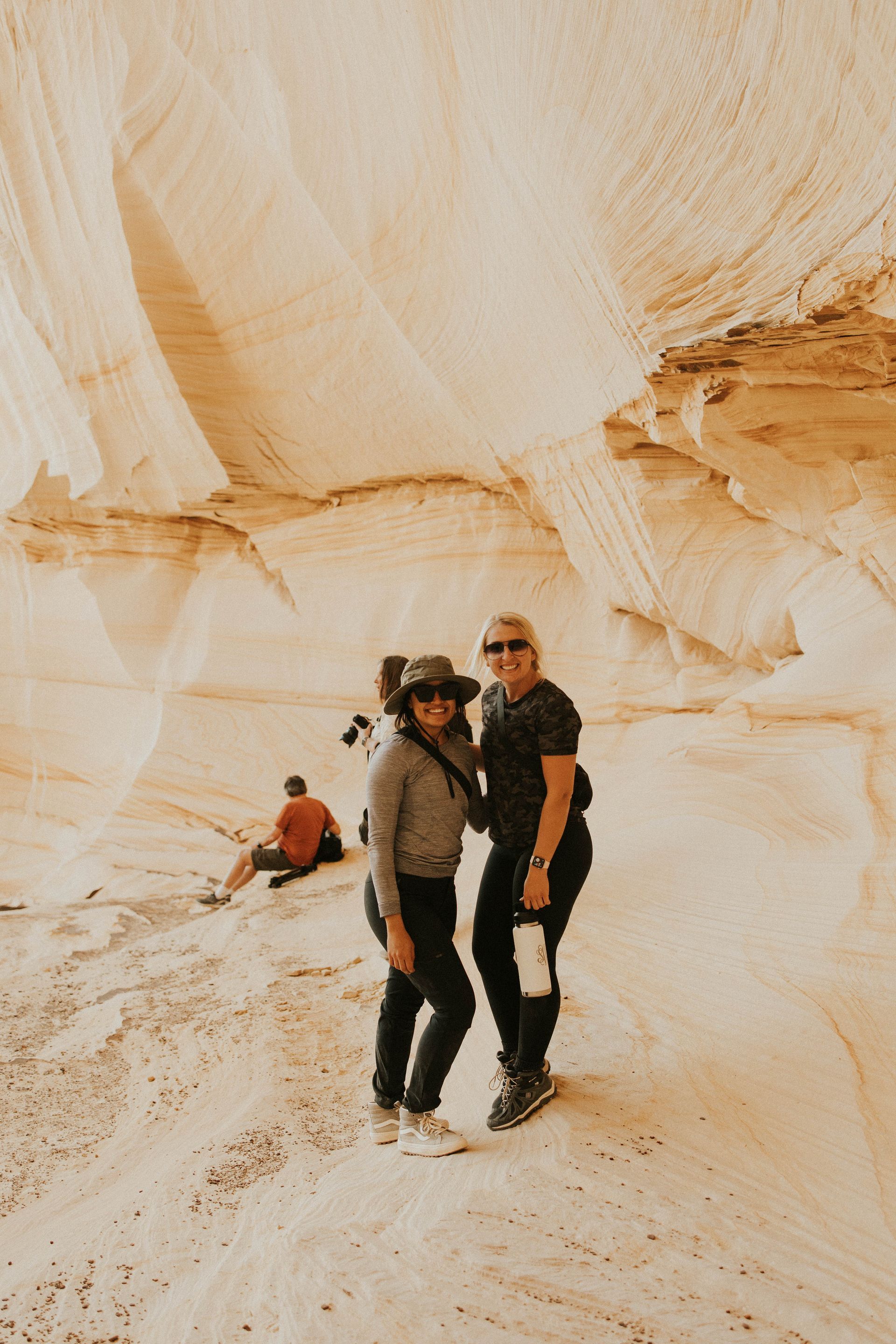 Two people stand smiling in a sandstone canyon while another person sits nearby in the background.